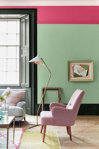 living room with green walls, pink painted above picture rail, painted skirtings, pink armchair, floor lamp, artwork, gray sofa, two rugs, vintage coffee table, wooden floor