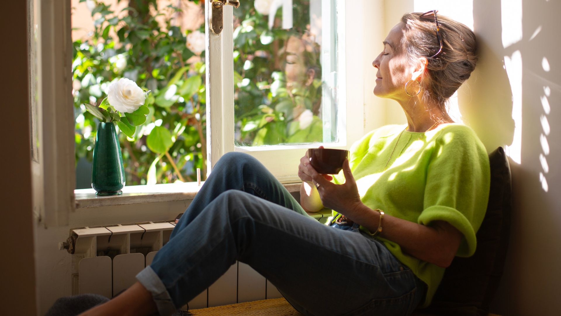 Woman drinking coffee sitting by window with sunshine coming through in the morning