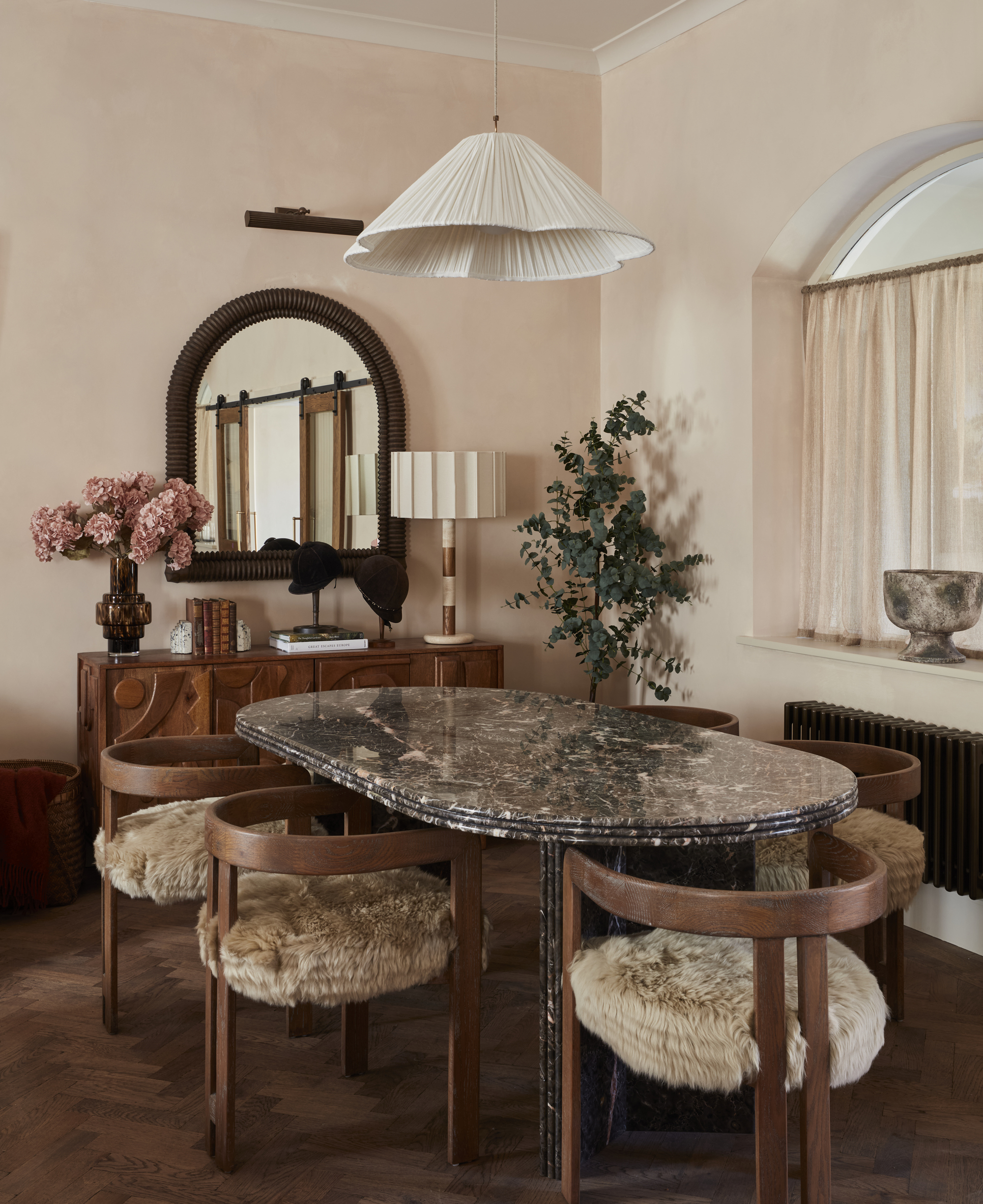 dining room with oval marble table, timber chairs with furry seats, plaster pink walls, arched mirror above a console with vase of flowers, lamp, and wall light