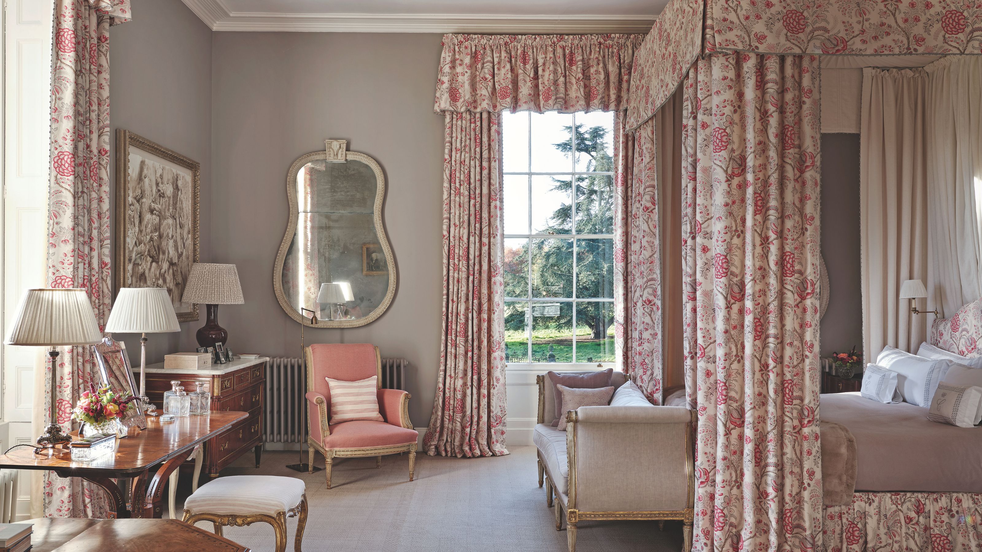 A grand, traditional bedroom featuring a four-poster bed with floral chintz fabric, a vintage pink armchair, and a large gilded mirror reflecting a soft grey wall.