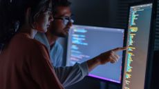 Male and female software developers in dimly lit room discussing memory safe languages while pointing at a vertically aligned desktop monitor.