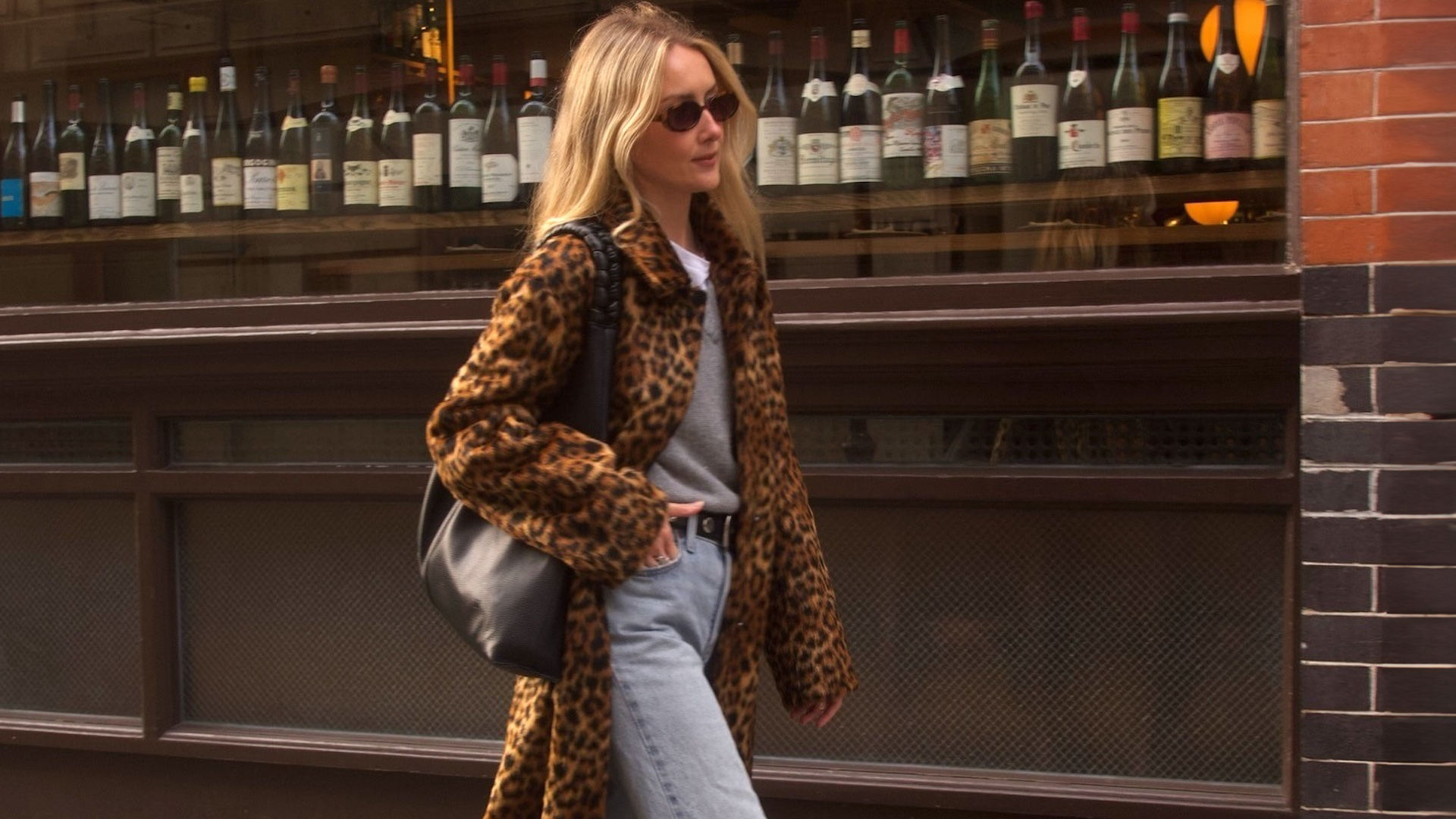 British style influencer Chloe Kath Butler walking on a London sidewalk wearing a leopard-print coat, gray V-neck sweater layered over a white T-shirt, studded black belt, black tote bag, and light-wash jeans