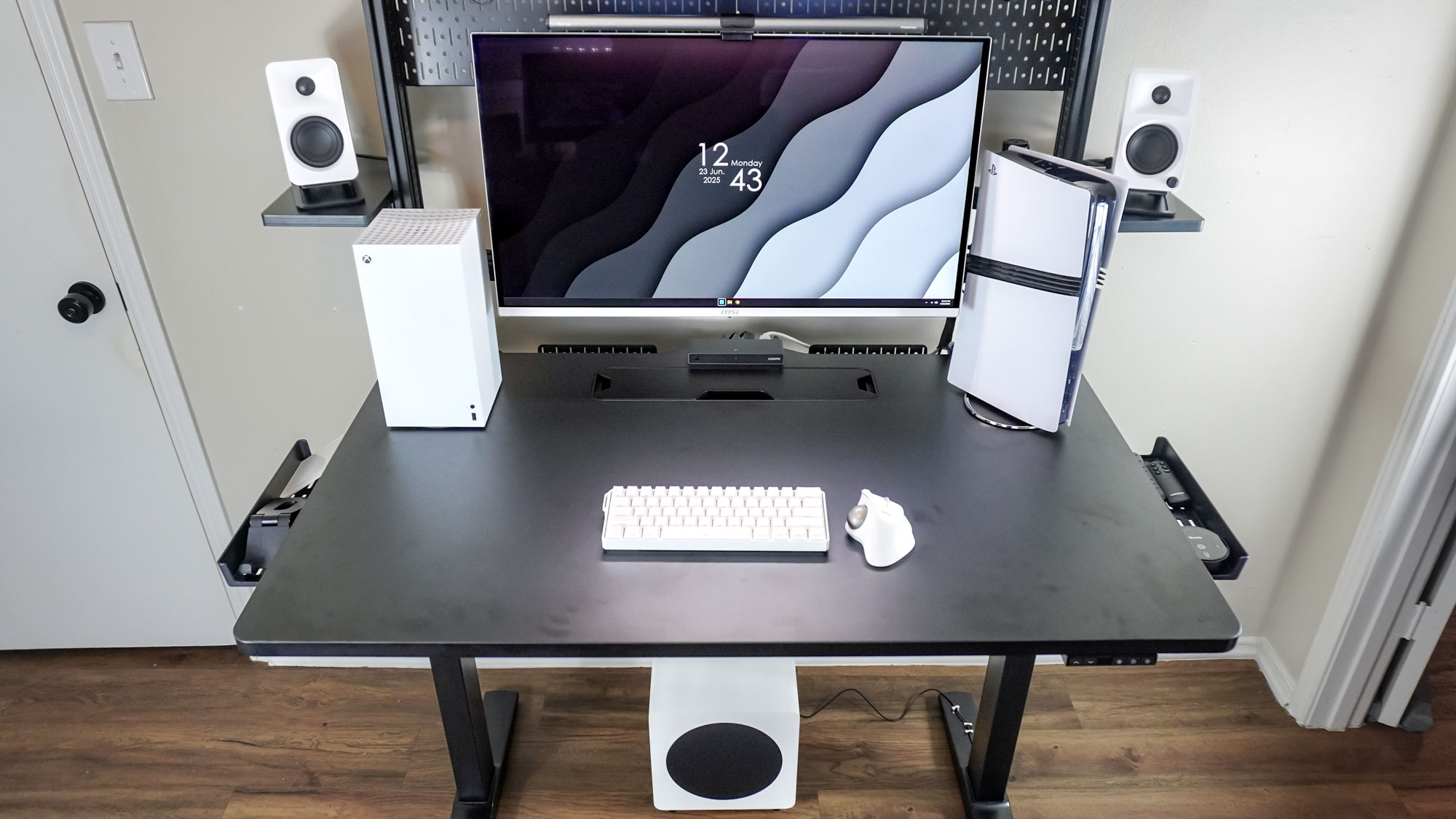 A wide shot of a black-and-white desk setup built around the Corsair Platform:4 Elevate Desk showing off the Kanto Ora bookshelf speakers up top and the Kanto SUB8 subwoofer underneath the desk