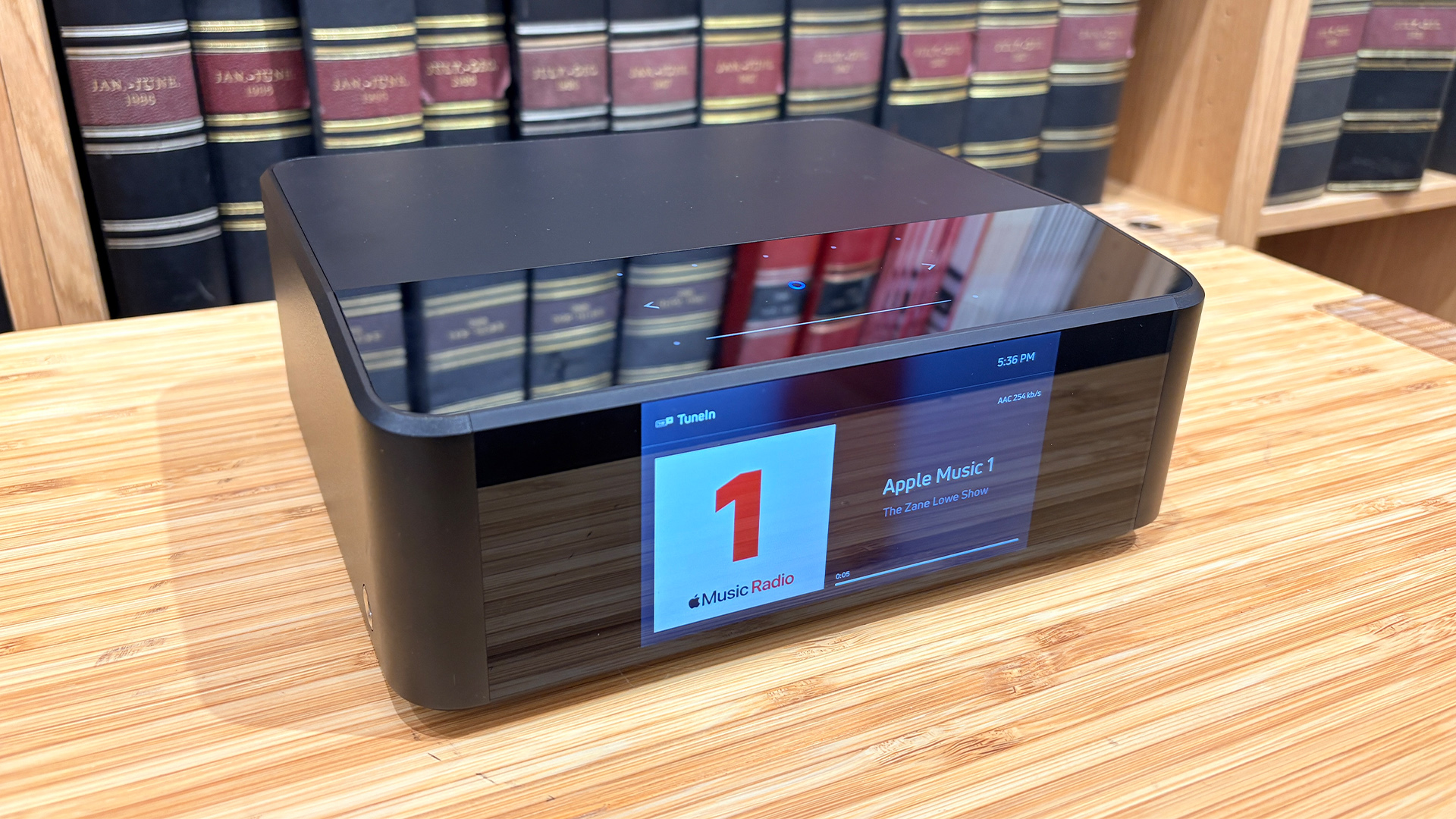 Bluesound Node Icon music streamer on wooden hi-fi rack in front of bookcase, on screen is Apple Music 1