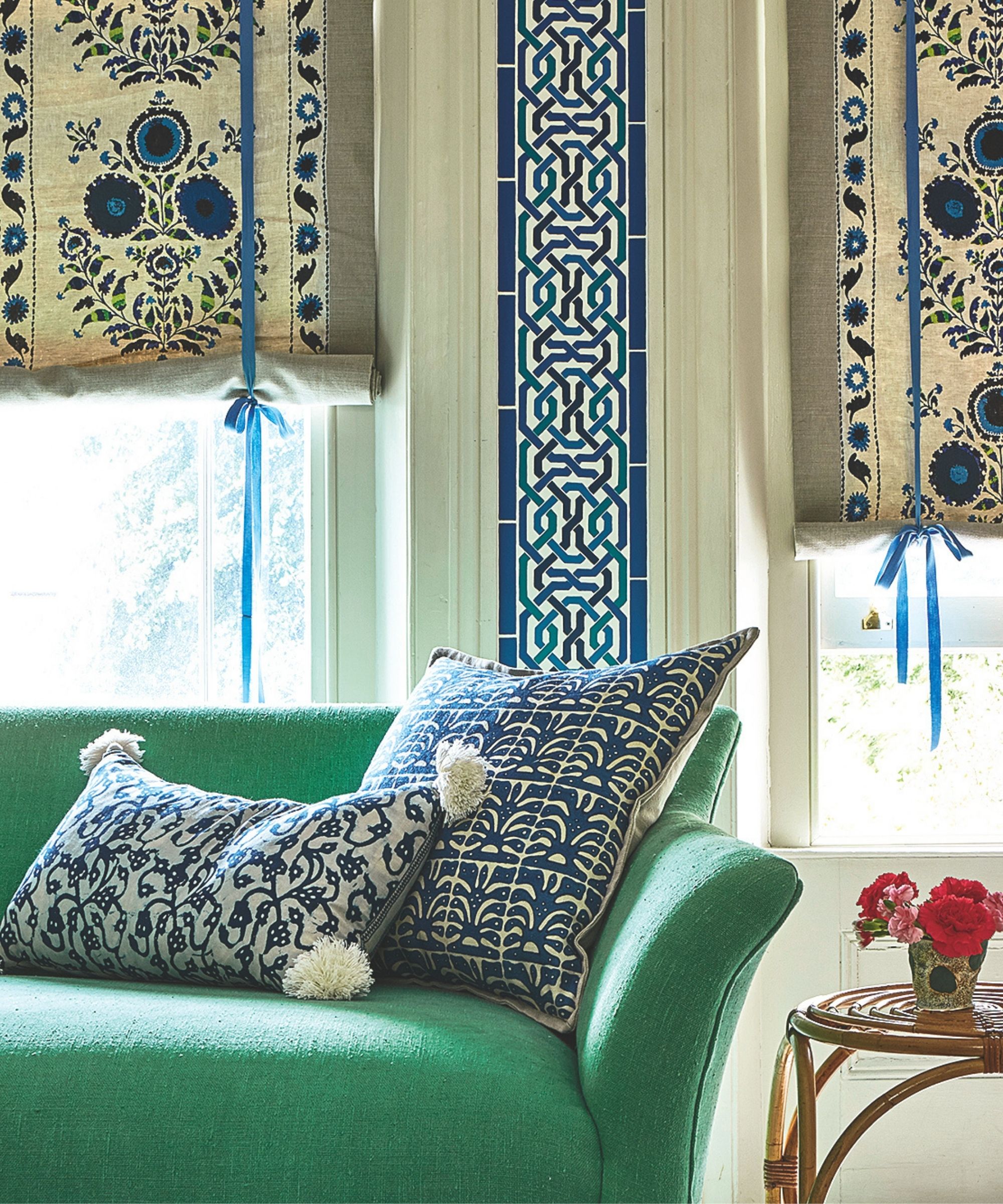 A green sofa with patterned blinds, patterned cushions, large windows, and a circular table with a small pot of red and pink flowers.