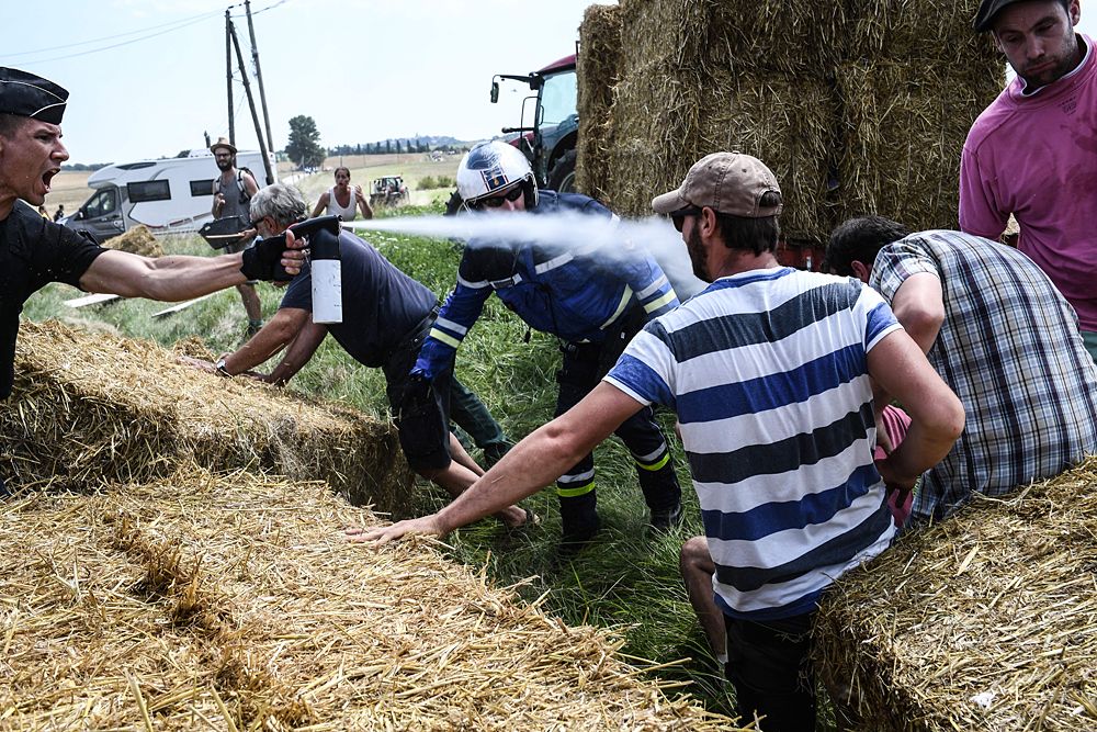 A gendarme (L) sprays tear gas at protesters (R, some hidden) as other gendarmes remove haystacks from the route, during a farmers' protest who attempted to block the stage's route