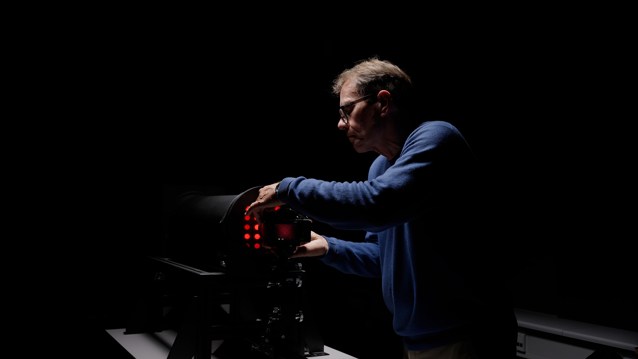 Man adjusting a camera or device with red lights in a dark room, illuminated by focused lighting