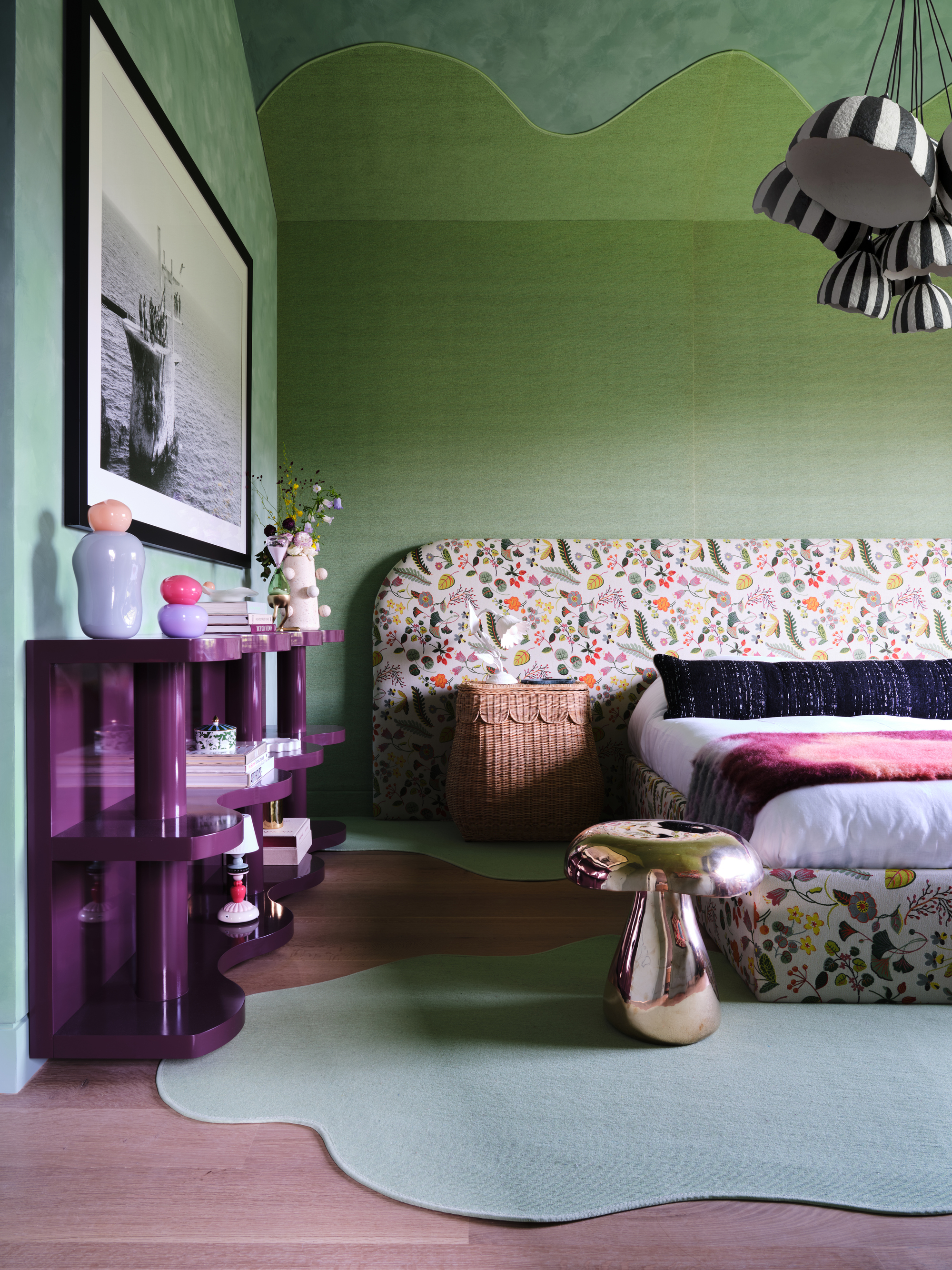 Image of a green bed room with a large floral patterned bed and a wicker side table. There is a purple shelving unit on the side and black and white striped lighting overhead.