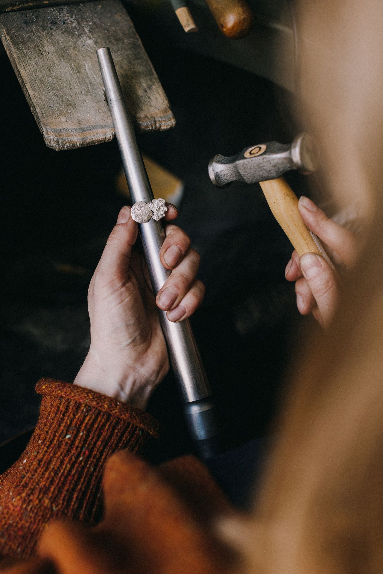 Image of two hands in a red sweater holding a hammer in one and a metal stick for crafting a ring.