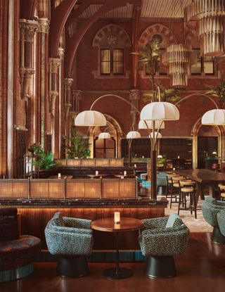 Inside the restaurant featuring different sections of tables and chairs, large overhanging lamps, tall ceiling. Colour scheme is predominantly dark wood brown, light brown and green pattern.