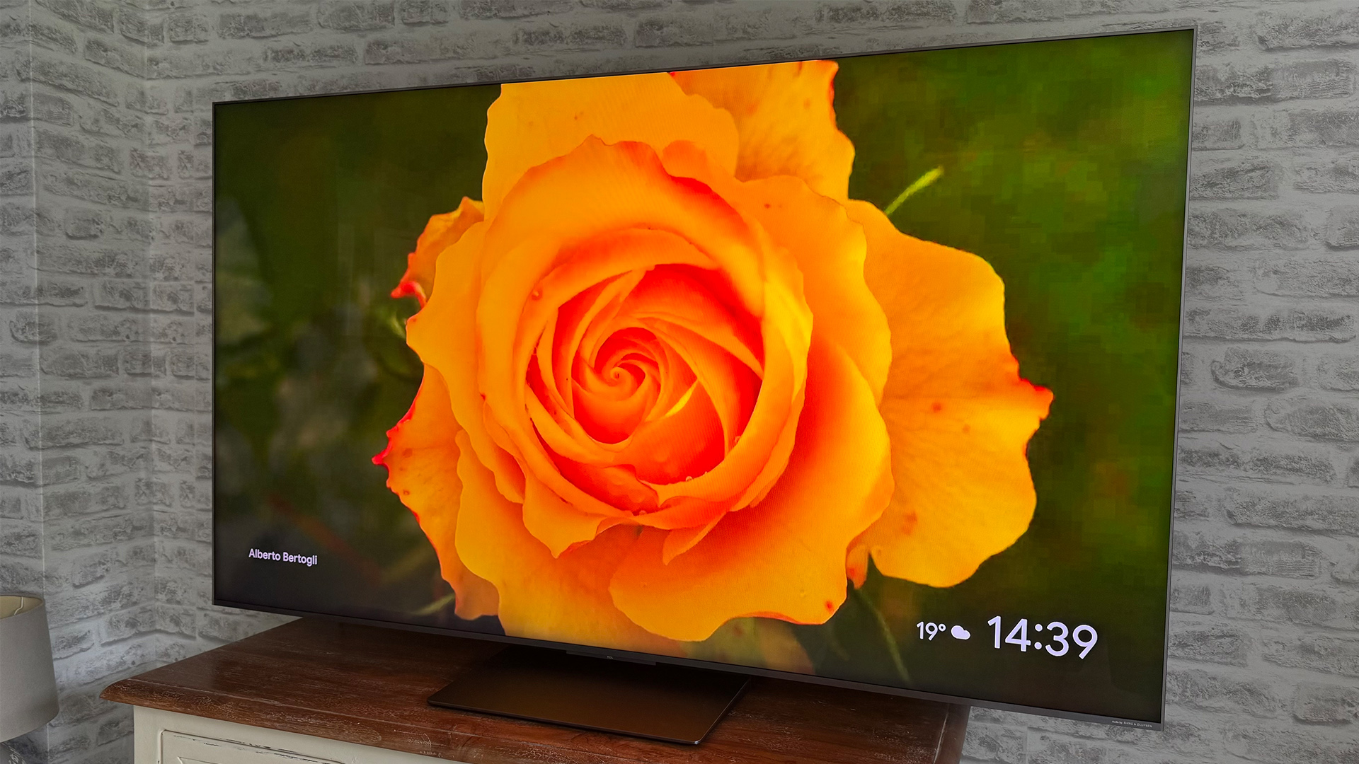 TCL 65C8K LCD TV on wooden cabinet in front of brick wall, on screen is orange rose