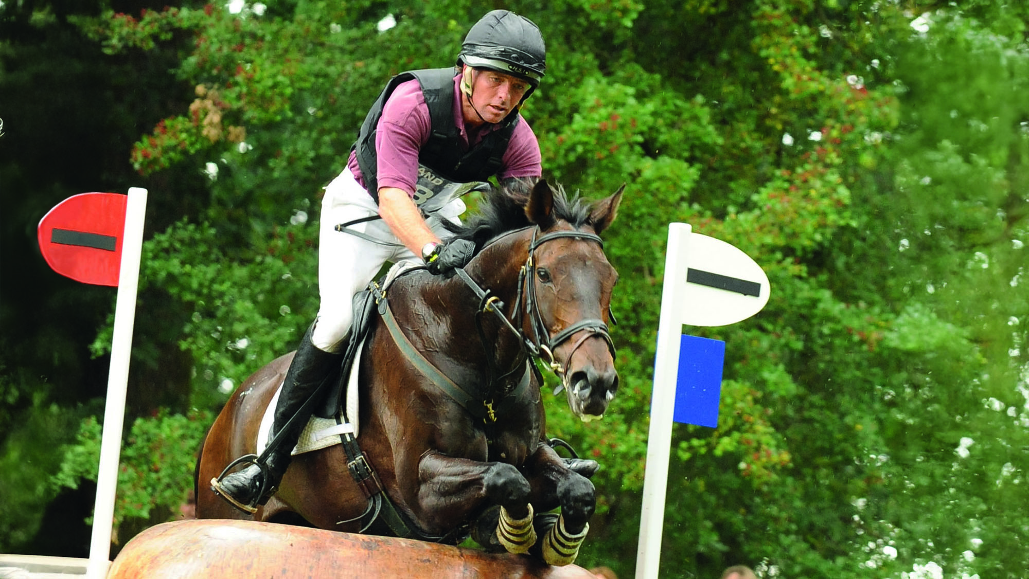 Dan Jocelyn, a rider in a dark red top with a black body protector, rides dark bay horse Silence over a a fence shaped like a mushroom at Burghley Horse Trials