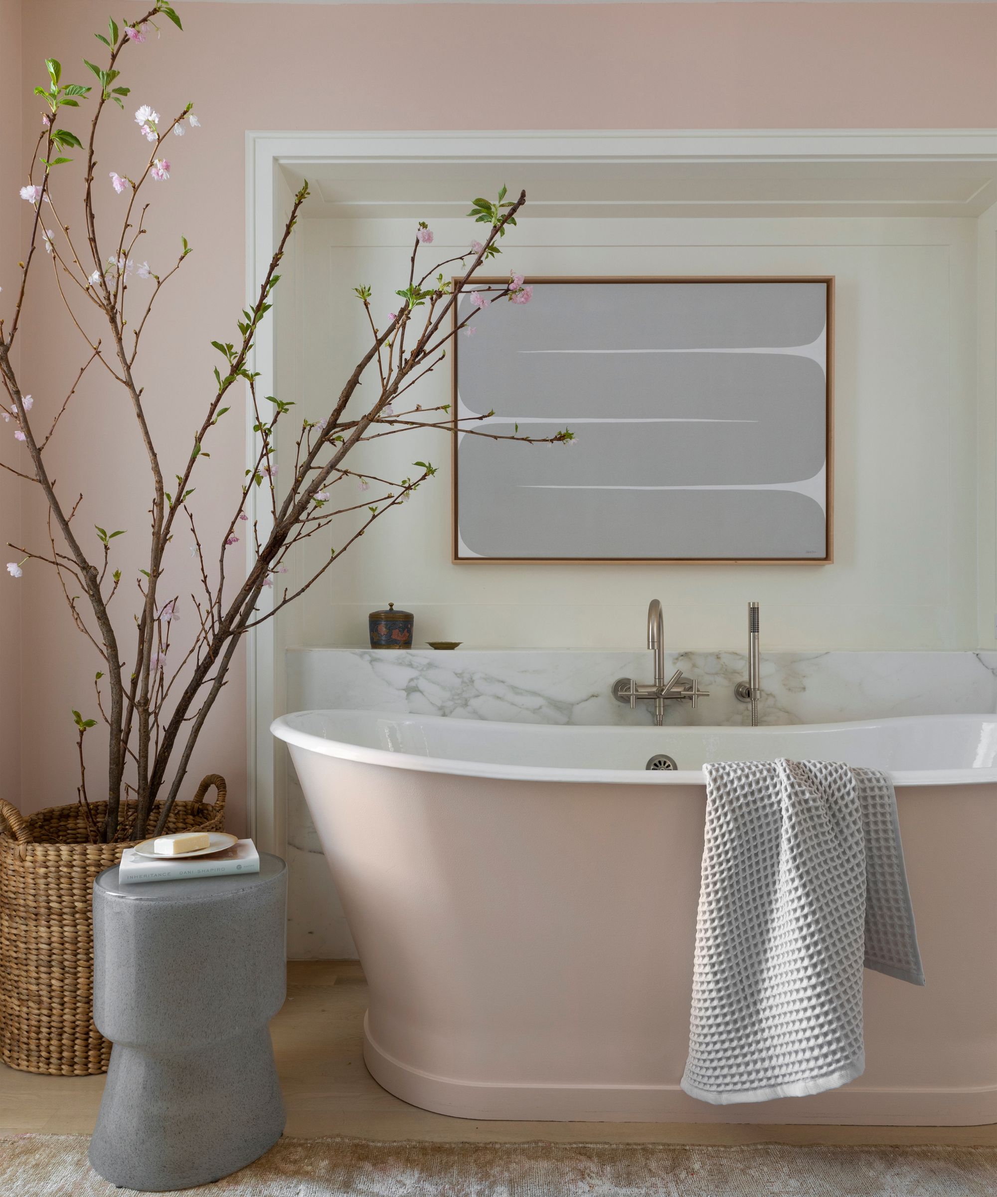 A calming bathroom with pale pink walls and a pale pink freestanding bath. A large potted plant in a wicker basket and a light gray abstract artwork on the wall.