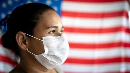 A woman with a face mask stands in front of an American flag