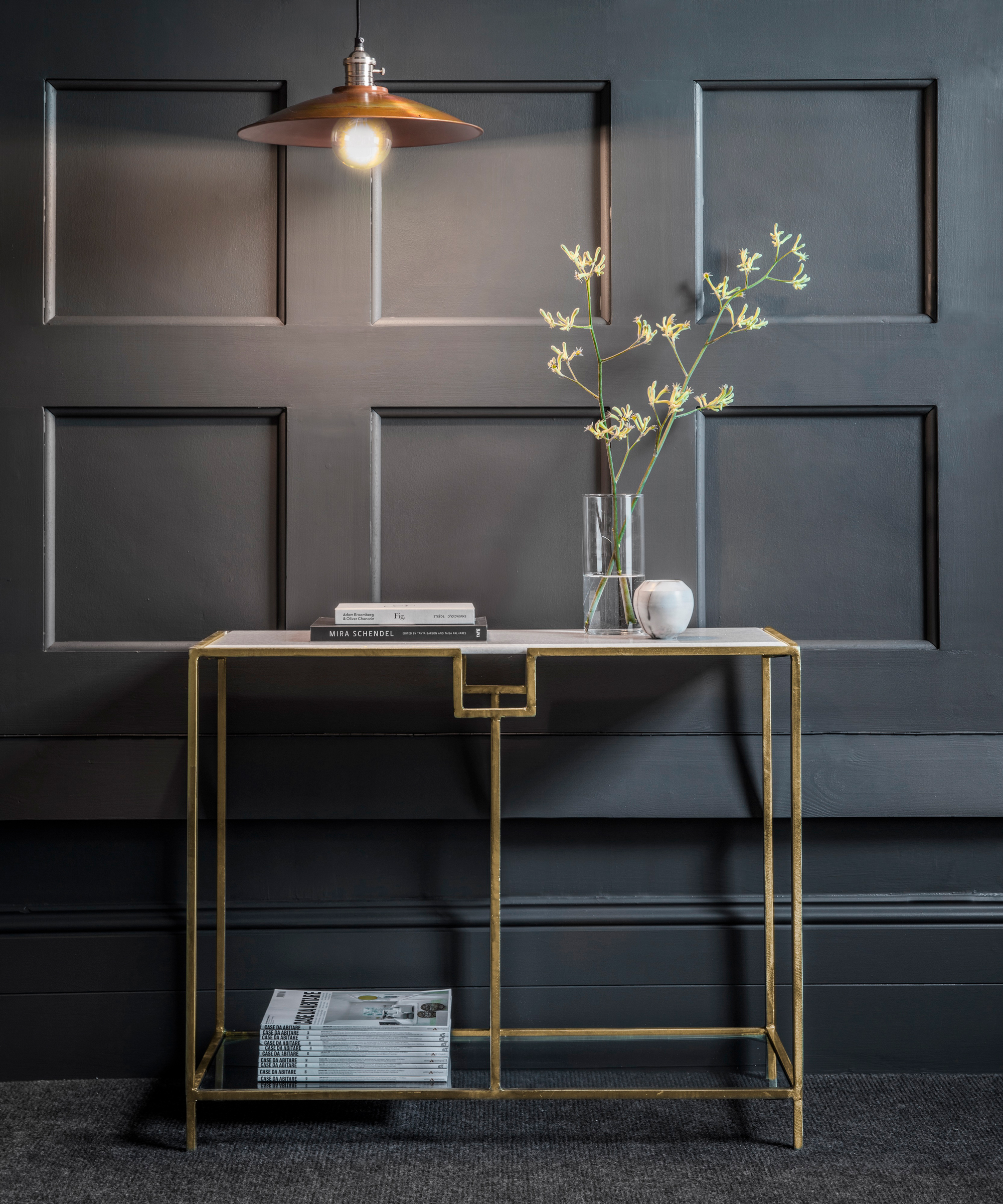 Dark, moody hallway with hat metallic pendant light, and slim brass console table with open storage space.