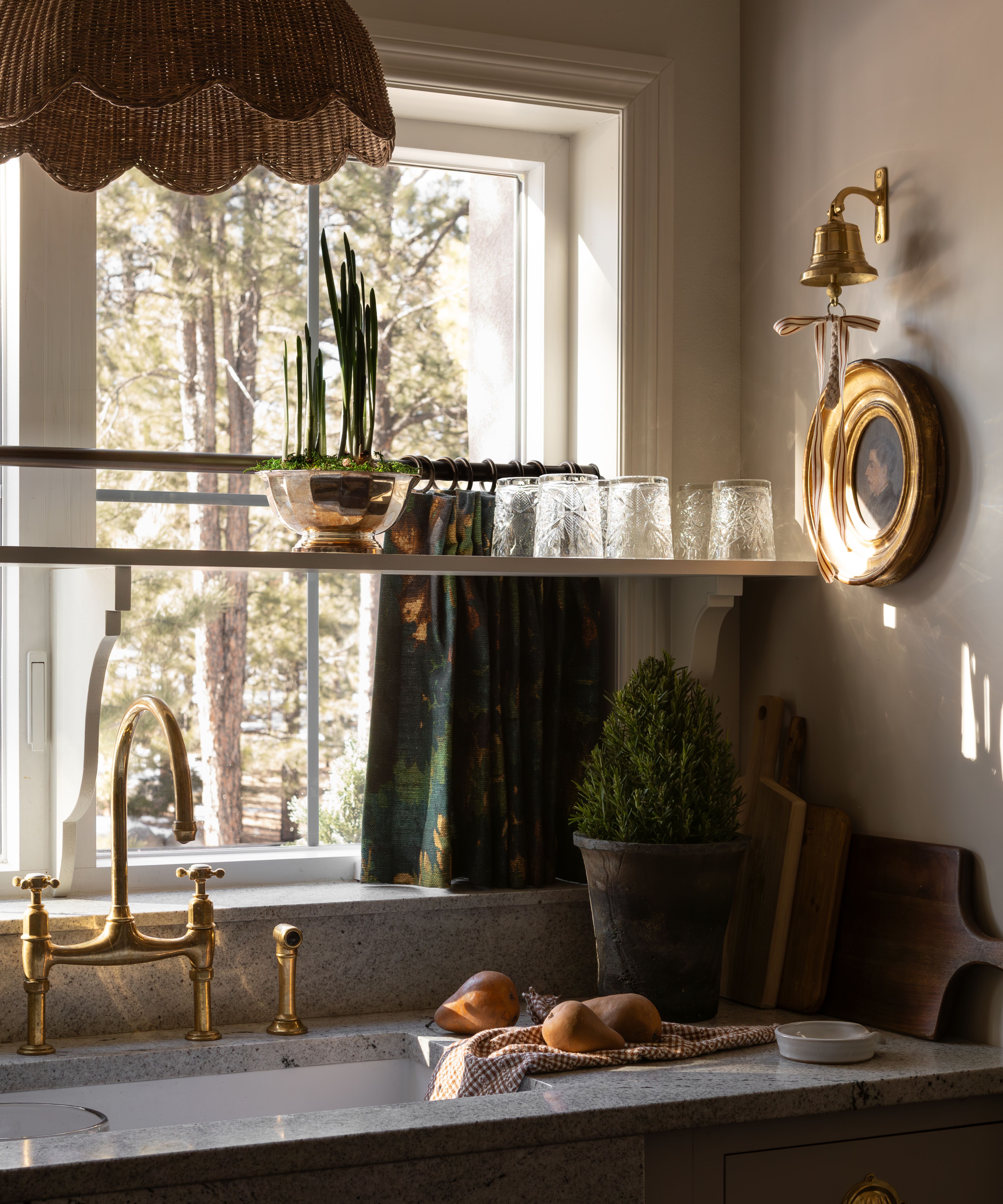 a small kitchen window and sink area with marble counters with a cafe curtain over the window, a shelf for glassware, a brass tap and on the wall, vintage art hangs with a brass bell