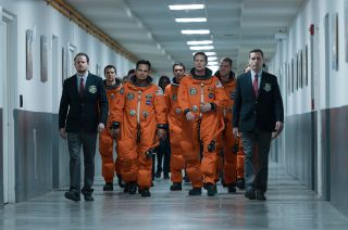 five astronauts in orange flight suits walk down a white-walled corridor.