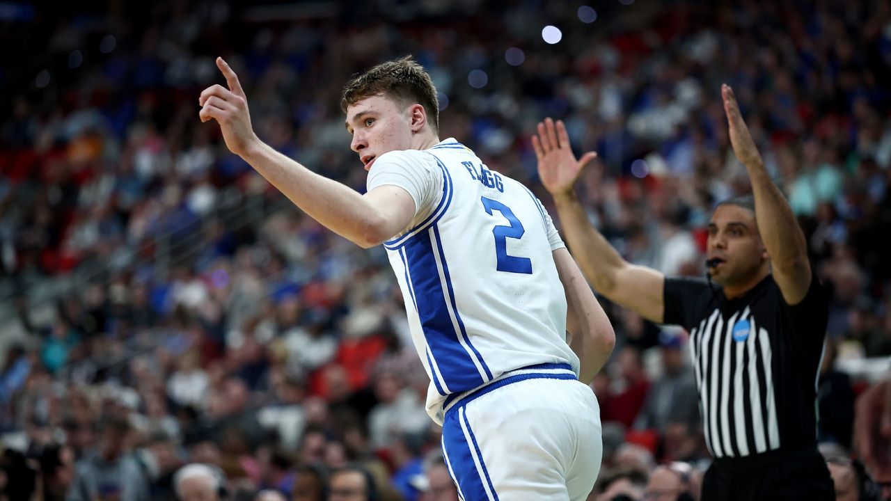 Cooper Flagg in his basketball uniform, in motion with his left arm raised and his index pointer in the air