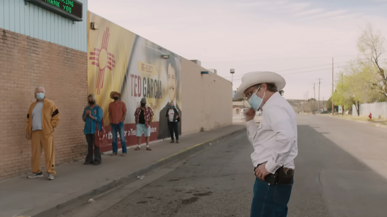 Joaquin Phoenix wearing a white cowboy hat and a medical mask looking at a line of people at a grocery store in Eddington