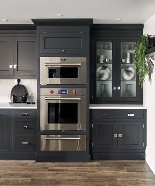 A dark gray kitchen with floor to ceiling cabinets. Two stainless steel ovens built in stacked on top of one another with a built-in proofing drawer beneath. White countertops.