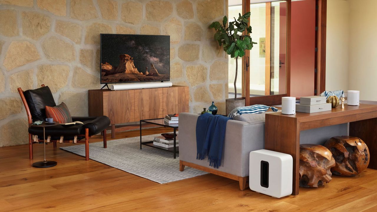 A photo of a living room with a TV and white Sonos Arc in the background and a white Sonos Sub next to a sofa in the foreground