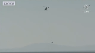 A helicopter holds the wrapped-up sample capsule. The two are seen mid-air with the sky in the background.