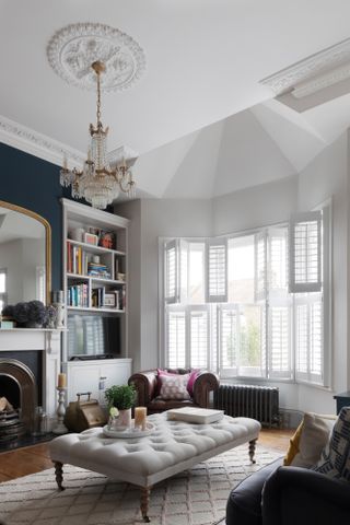 Pippa Jones house: living room with big bay window, white shutters, white walls and dark blue feature wall, beige buttoned ottoman coffee table and pink and white rug