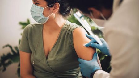 A young woman in a surgical mask sit in a doctor's office as a doctor cleans her arm for a vaccination