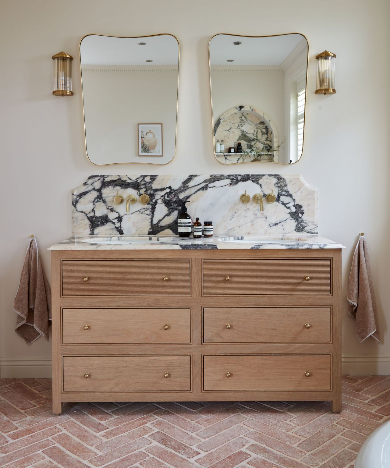 A warm white bathroom with terracotta floor tiles, gold hardware, and a wooden vanity topped with marble