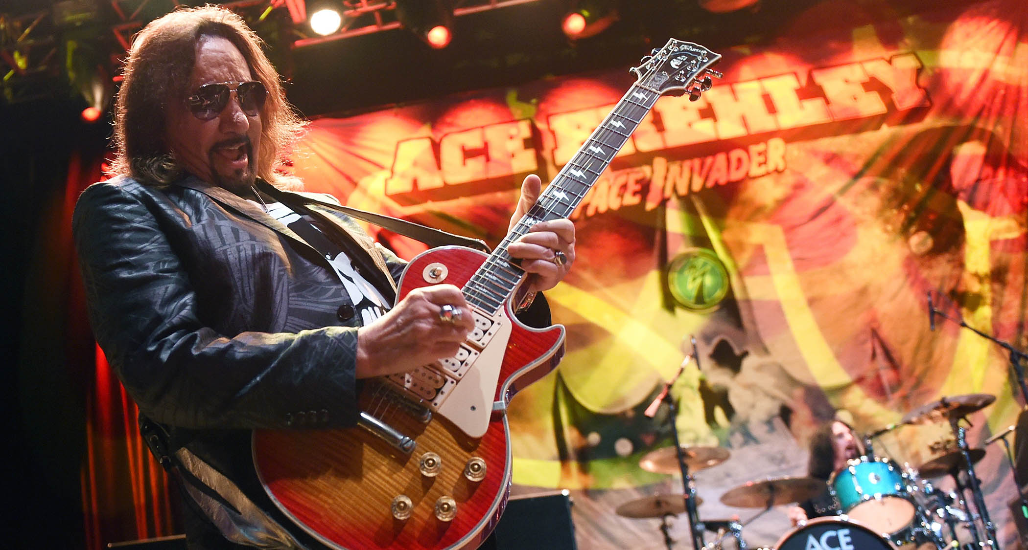 A close-up live shot of Ace Frehley performing with his trademark sunburst triple-humbucker Les Paul