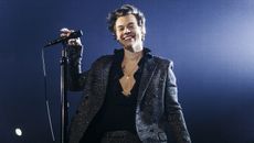 harry styles on stage holding a microphone smiling while wearing a sparkly suit