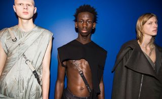 Three male models wearing looks from the Rick Owens collection. One model is wearing a grey multi-layered piece with chain and straps. Next to him is a model wearing dark bottoms and a black top that exposes the chest and arms and features a chain and strap. The third model is wearing a grey piece with chain and strap and a dark grey coat over the top. They are standing in front of a blue wall