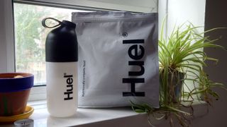 A Huel shaker next to a bag of Huel powder, a potted plant and a cactus.