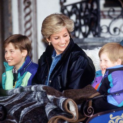 Princess Diana, Prince William and Prince Harry sitting in a sleigh with blankets and wearing bright ski coats in the 1990s