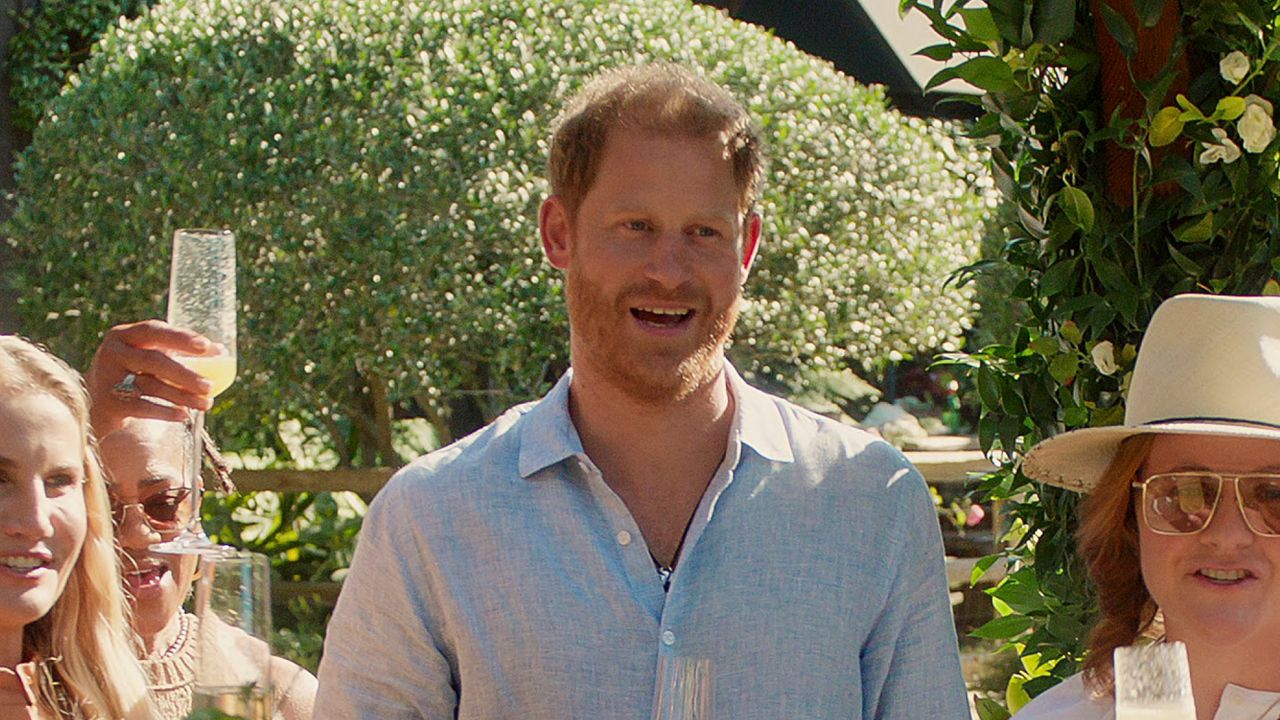 Prince Harry has red hair and beard and wears a light blue linen shirt while holding a champagne glass in With Love, Meghan