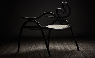 Side view of a black chair by Studio Swine featuring a curved back that resembles the shape of a snake and a woven seat in a contrasting colour. The chair is pictured in a space with a dark wall and carpet