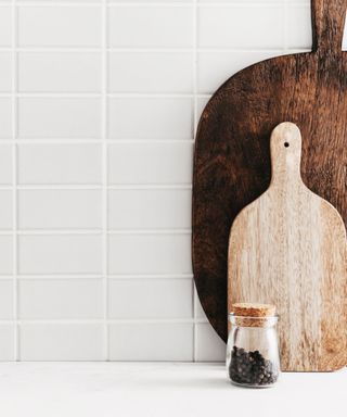 White kitchen tiles with a white countertop with two wooden chopping boards - one light brown and one dark brown - with a glass pot of pepper in front