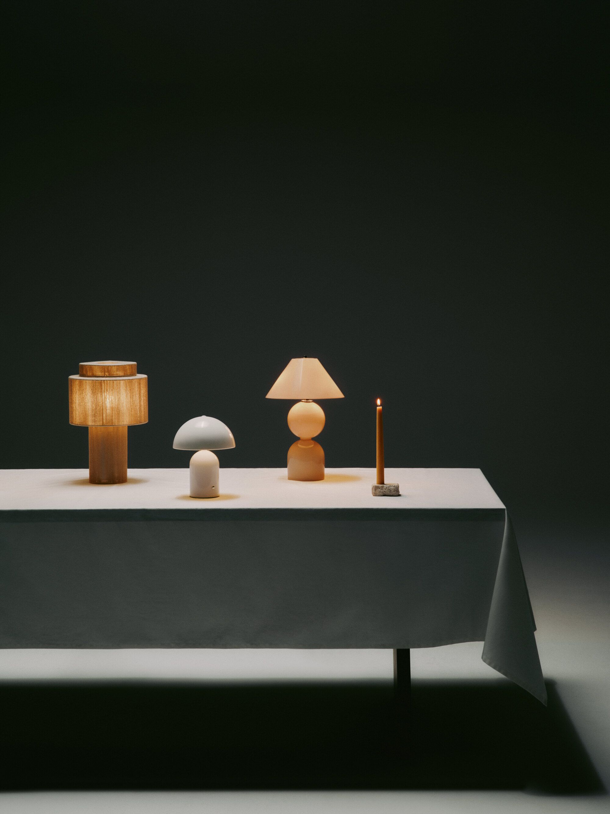 Four modern table lamps arranged on a long table with a white tablecloth on it in a white and gray showroom.