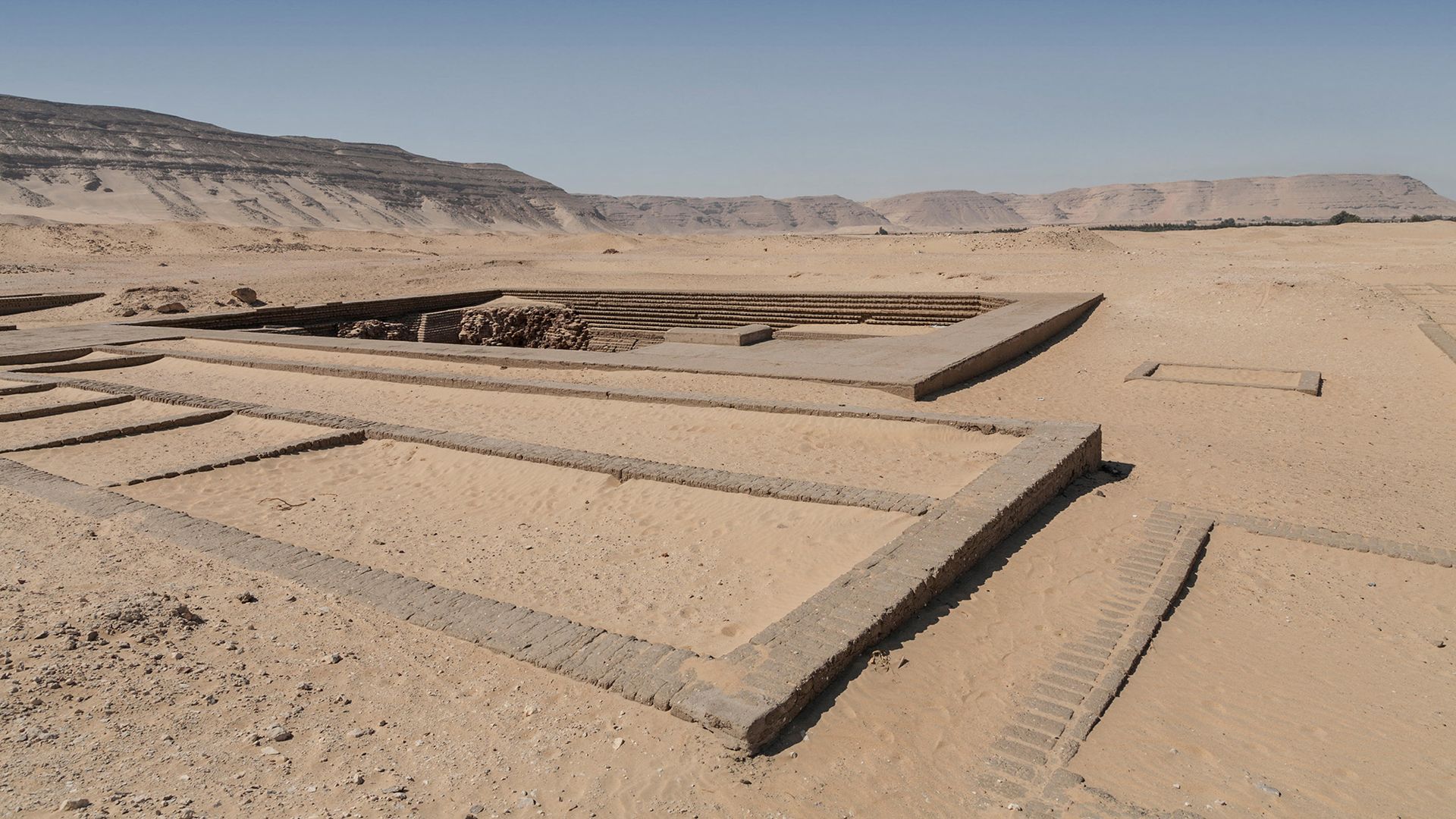 A series of square areas are seen in the desert, with a larger tomb in the background