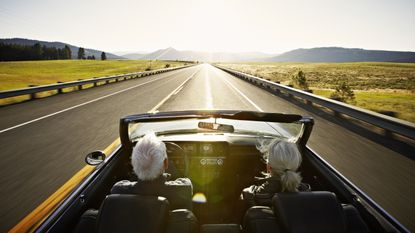 Two older adults drive toward the sun and mountains in an open convertible.
