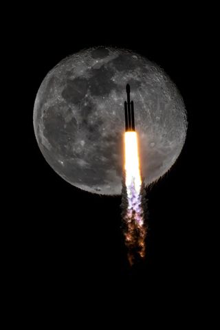a silhouetted triple booster rocket passes in front of a full moon as fire spews from its engines.
