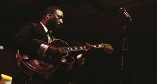 Jazz guitar legend Wes Montgomery plays his Gibson hollowbody on a dimly lit stage. He wears a black suit. Montgomery is exactly the kind of guitarist that gets sampled for lo-fi tracks.