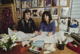 David and Elizabeth Emanuel sitting at a table covered in sketches and magazines and looking at the camera