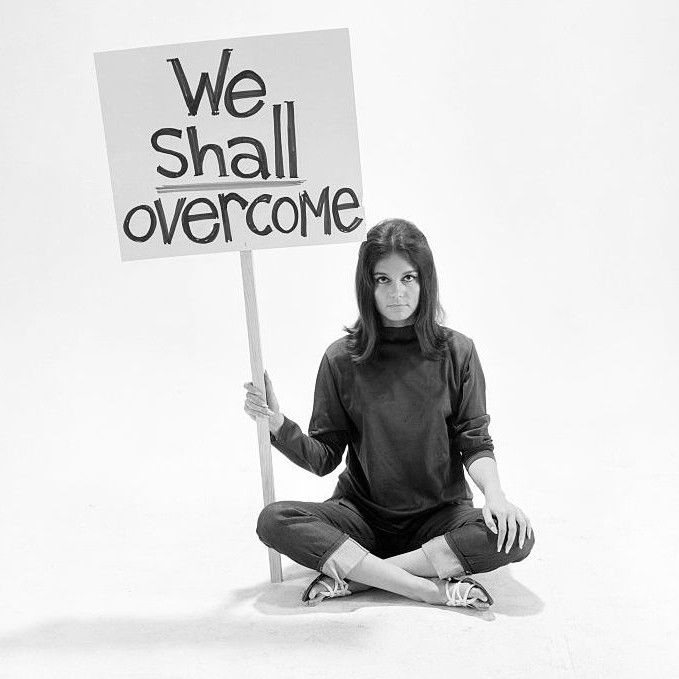 A woman in 1960s clothes holding a placard that says "we shall overcome"