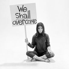 A woman in 1960s clothes holding a placard that says "we shall overcome"
