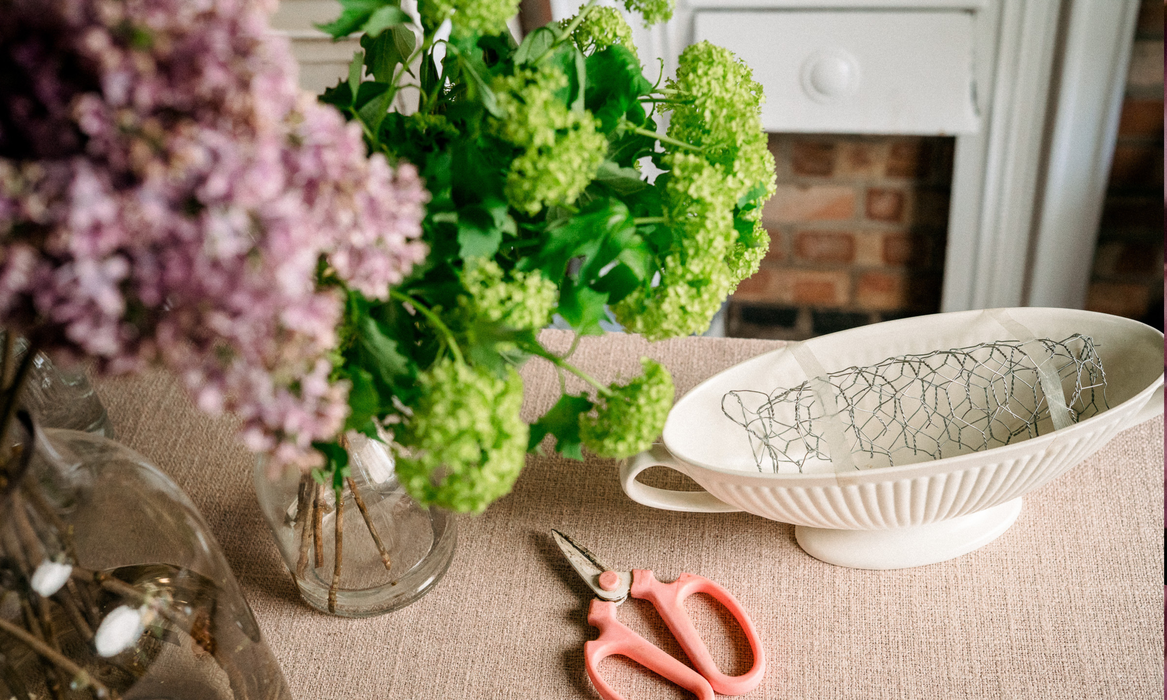 Pink scissors next to white ceramic boat vase filled with chicken wire and green viburnum opulus flowers