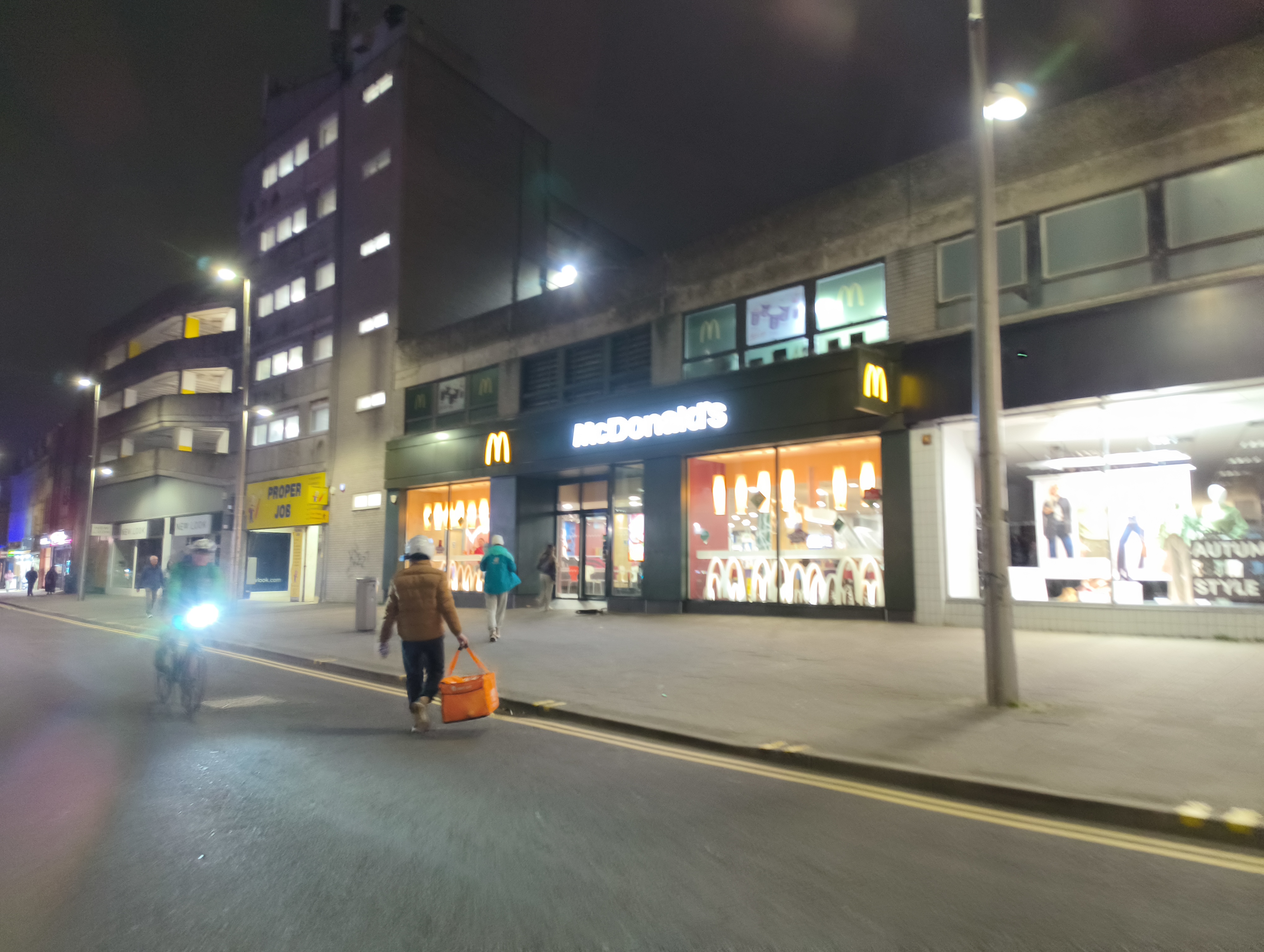 A nighttime street view of a brightly lit McDonald's restaurant with orange and black exterior signs, as a person walks toward the entrance and a cyclist passes on the left.