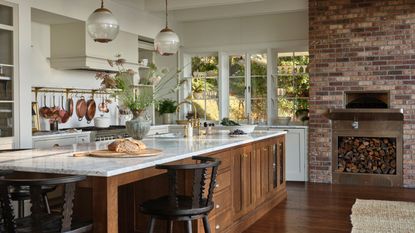 A spacious, sunlit kitchen featuring a large oak island with a marble countertop, black spindle stools, and vintage-style glass pendant lights