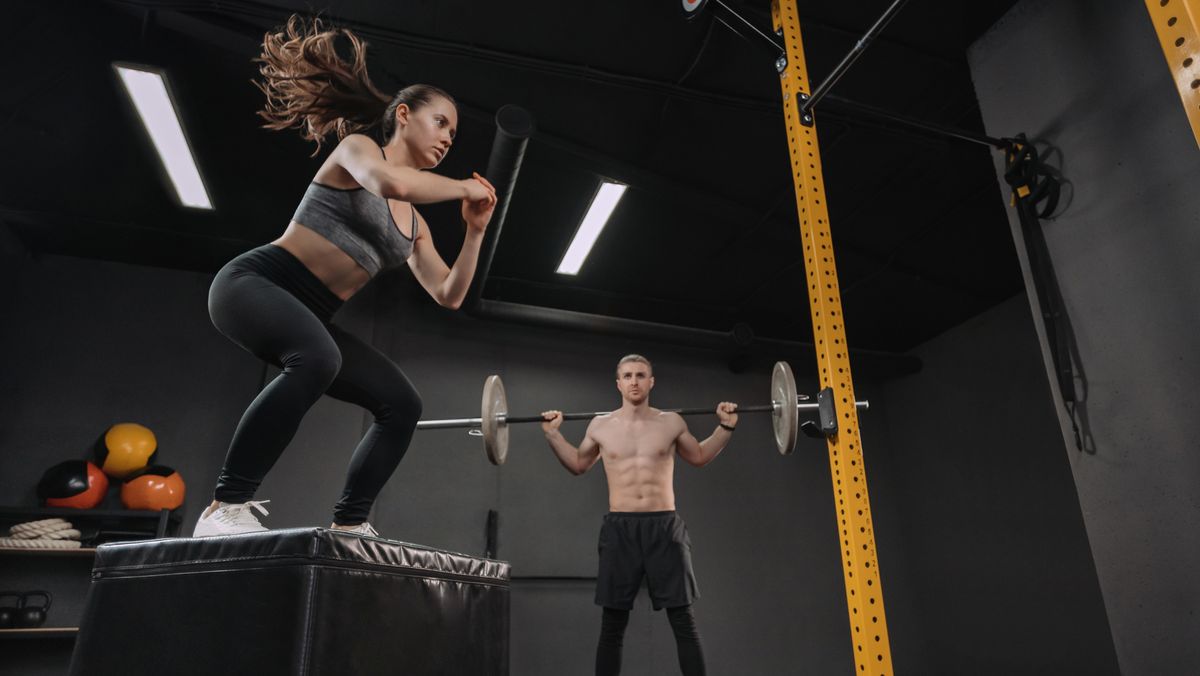 Woman performs box jump exercise in gym, man holds barbell across the back of his shoulders in background