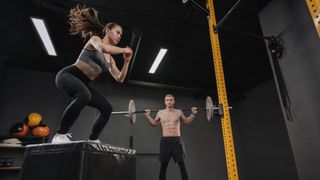 Woman performs box jump exercise in gym, man holds barbell across the back of his shoulders in background